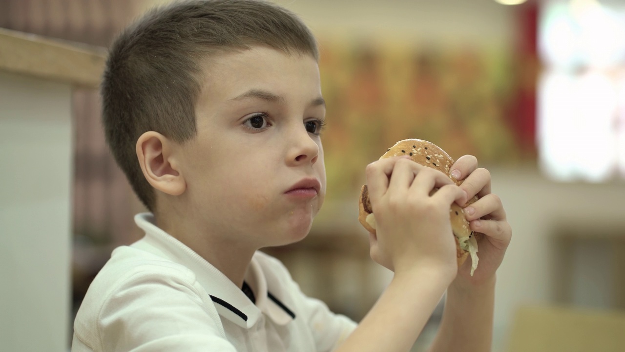 小学生坐在咖啡馆里吃汉堡包。男孩手里拿着一个汉堡吃了起来。视频下载