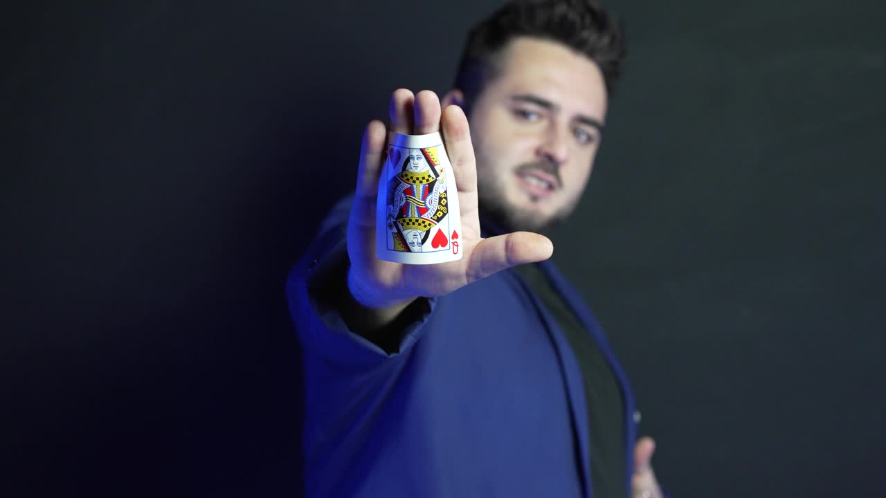模糊的视频肖像，一个年轻的魔术师在黑暗中显示在他的手掌隐藏的卡，同时看着摄像机，并揭示它。显示的卡片已聚焦。视频下载