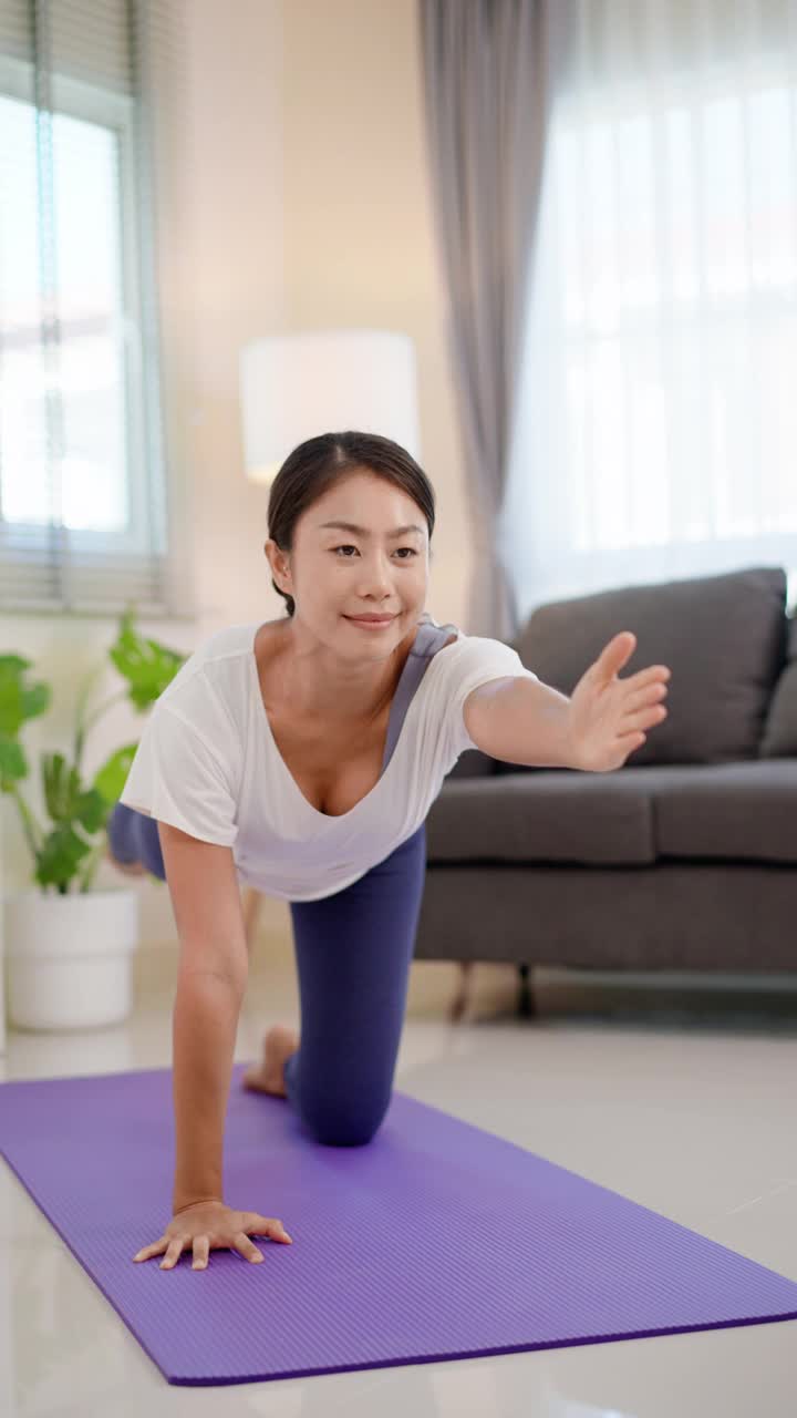 年轻活跃的运动苗条的亚洲女性做瑜伽锻炼，伸展一条腿到背部和抬起她的手臂伸展向前，以提高肌肉灵活性在客厅在家健身。视频下载