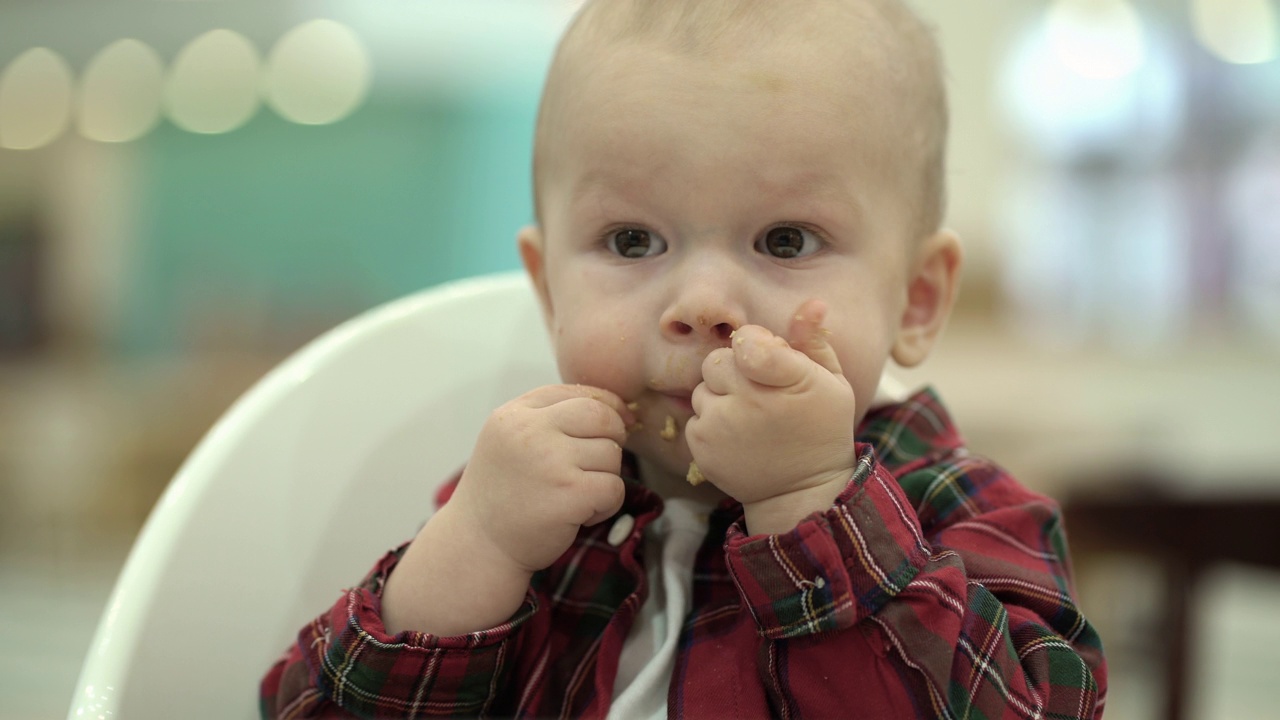 一个小孩坐在儿童椅上，用手吃着碎饼干。一个被美味的饼干弄脏了脸的男孩的肖像。视频下载