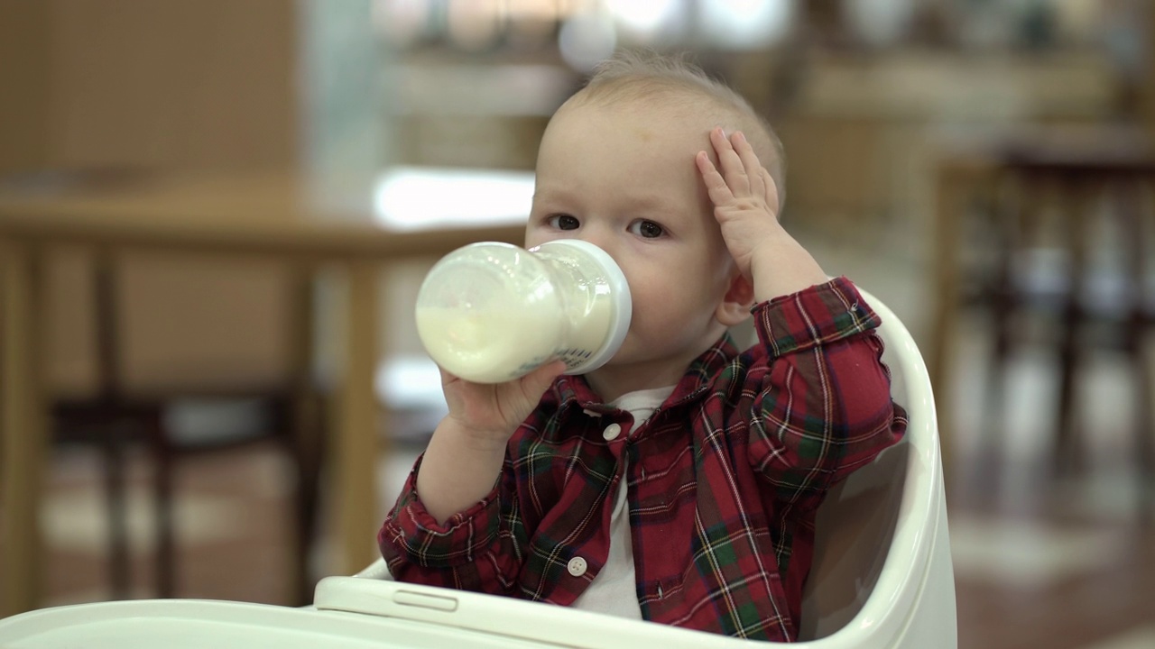 一个一岁大的男孩坐在婴儿椅上，手里拿着一个装有婴儿配方奶粉的奶瓶。吃婴儿食品的孩子。视频下载