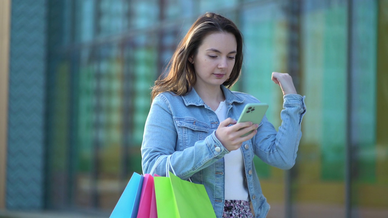 从商店拿着彩色纸袋的女人走在街上，用她的手机与人交流视频下载
