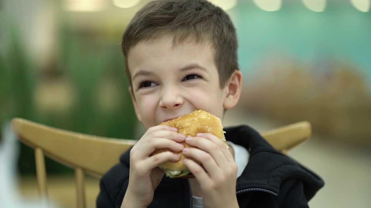 一个疲倦悲伤的七岁男孩放学后在美食广场吃汉堡。穿着运动衫的孩子正在享用多汁的汉堡。视频下载