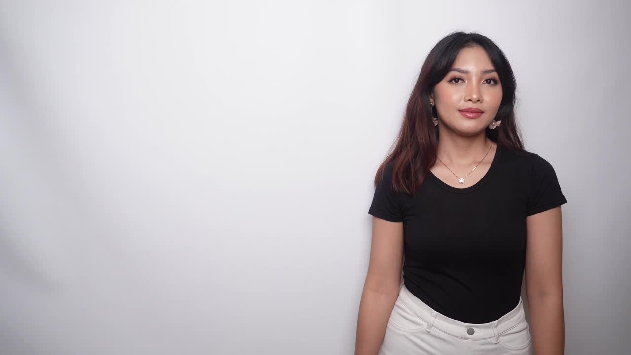 一个穿着黑色衬衫的兴奋的亚洲女人，指着她旁边被白色背景隔开的复印空间视频下载