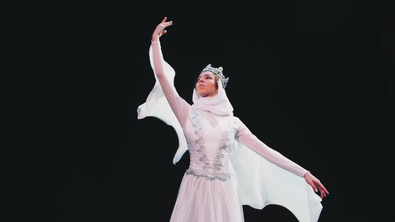 在黑色背景的剧场舞台上，身穿美丽的白色套装、头戴皇冠的芭蕾舞女演员正在表演美丽的芭蕾元素。风吹着她的衣服。一只手举起来。美丽的姿势。视频素材
