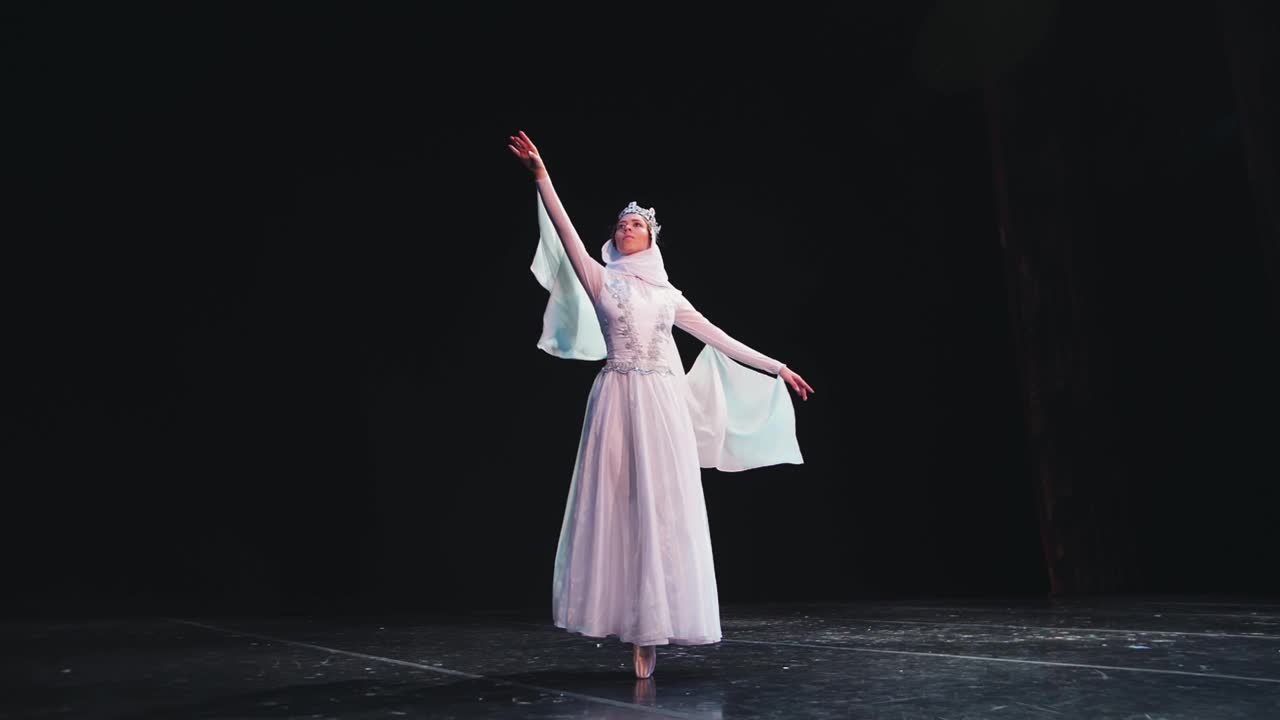一位优雅、勤劳的芭蕾舞女演员头戴皇冠，在黑色背景上摆出美丽的姿势。风吹着她美丽的白色套装。视频下载