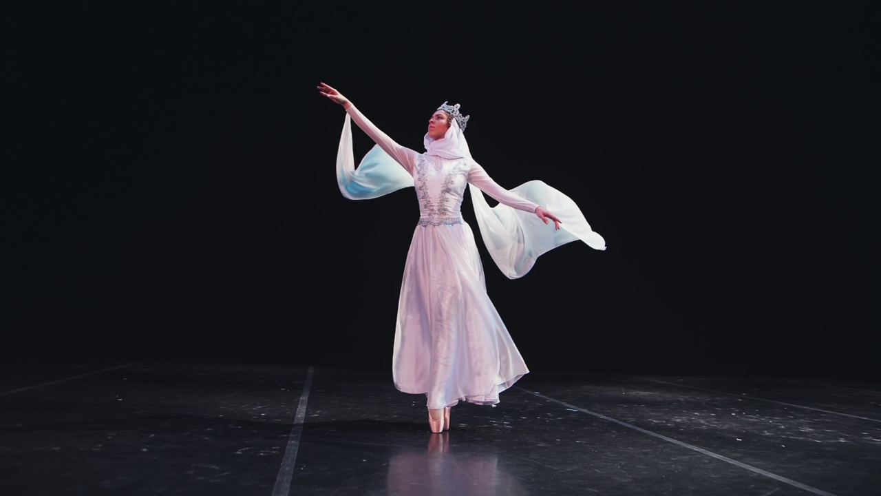 身穿美丽的白色套装、头戴皇冠的芭蕾舞女演员在黑色背景的剧场舞台上摆出了美丽的芭蕾舞姿势。风吹着她的衣服。一只手举起来。缓慢的运动。视频素材