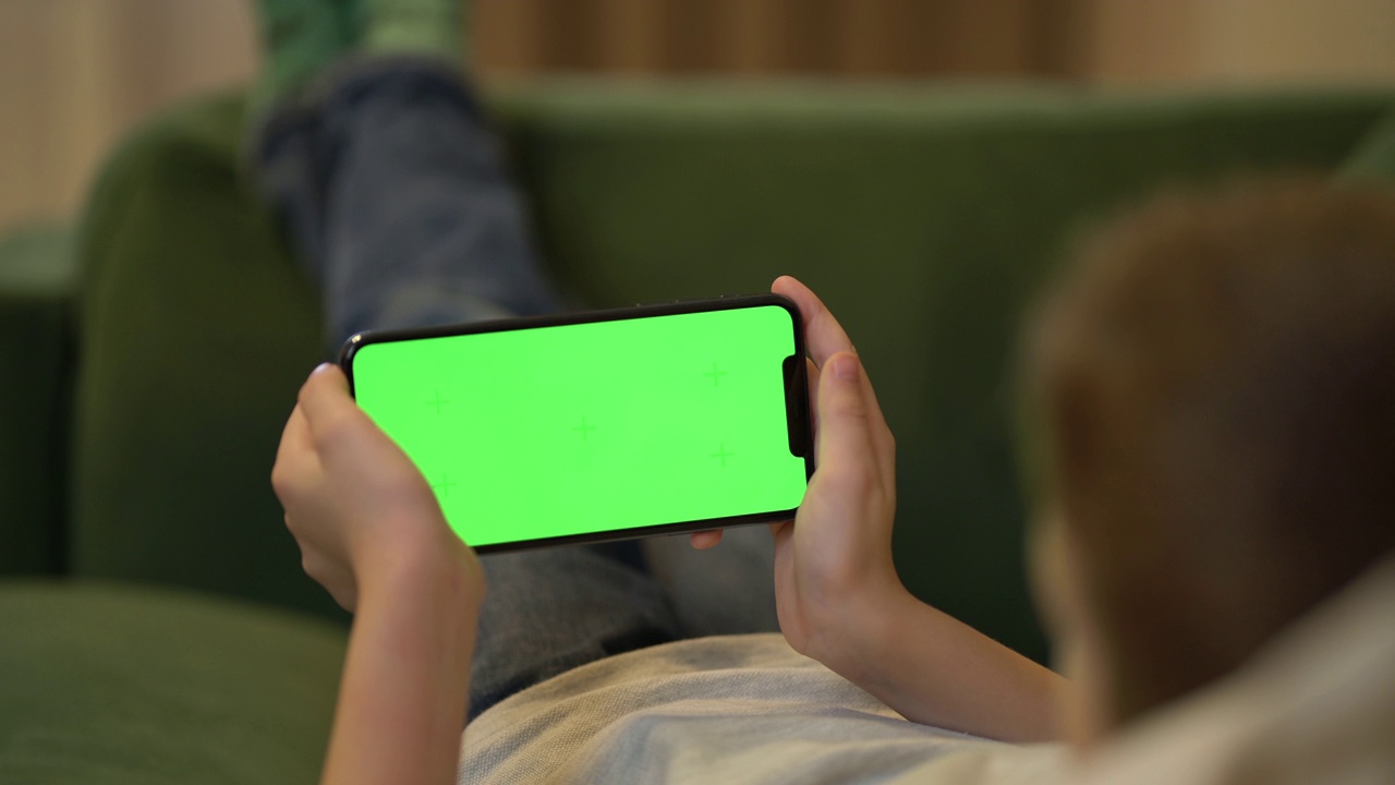 男孩躺在沙发上使用绿屏智能手机的后视图。小学生手里的色度键手机视频下载