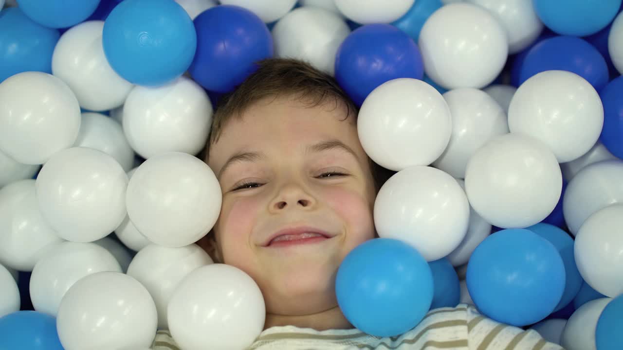 一个男孩躺在泳池里的一堆塑料球里。视频下载