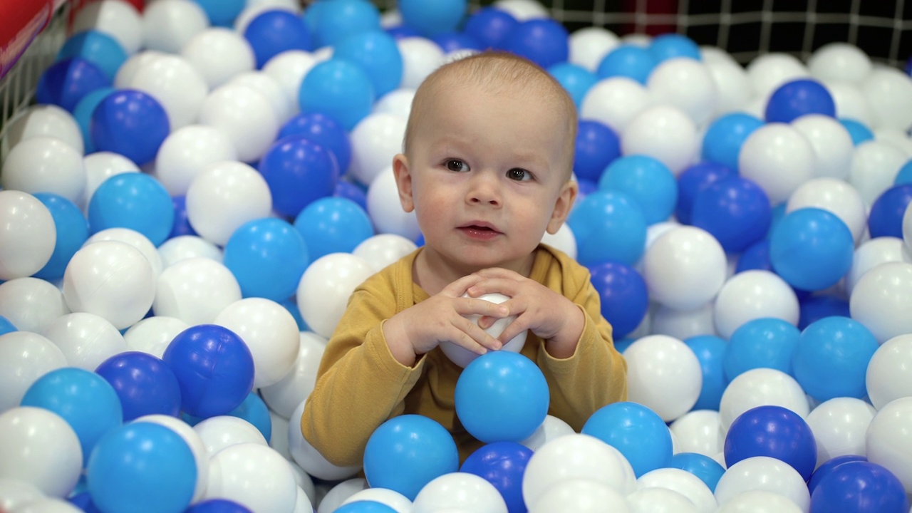 一个一岁大的男孩坐在游泳池里，玩着五颜六色的塑料球。视频下载