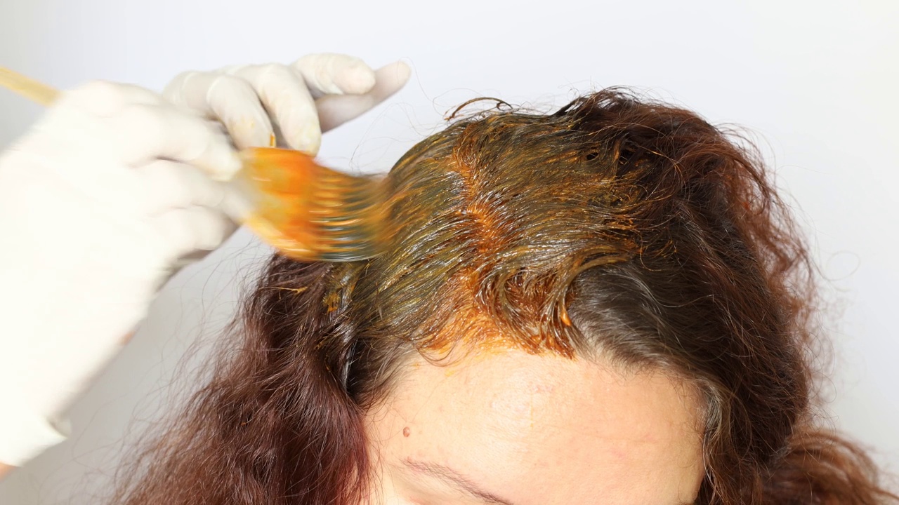理发师戴着白手套，将橙色染发剂涂抹在客户深棕色长发的发根上。视频下载