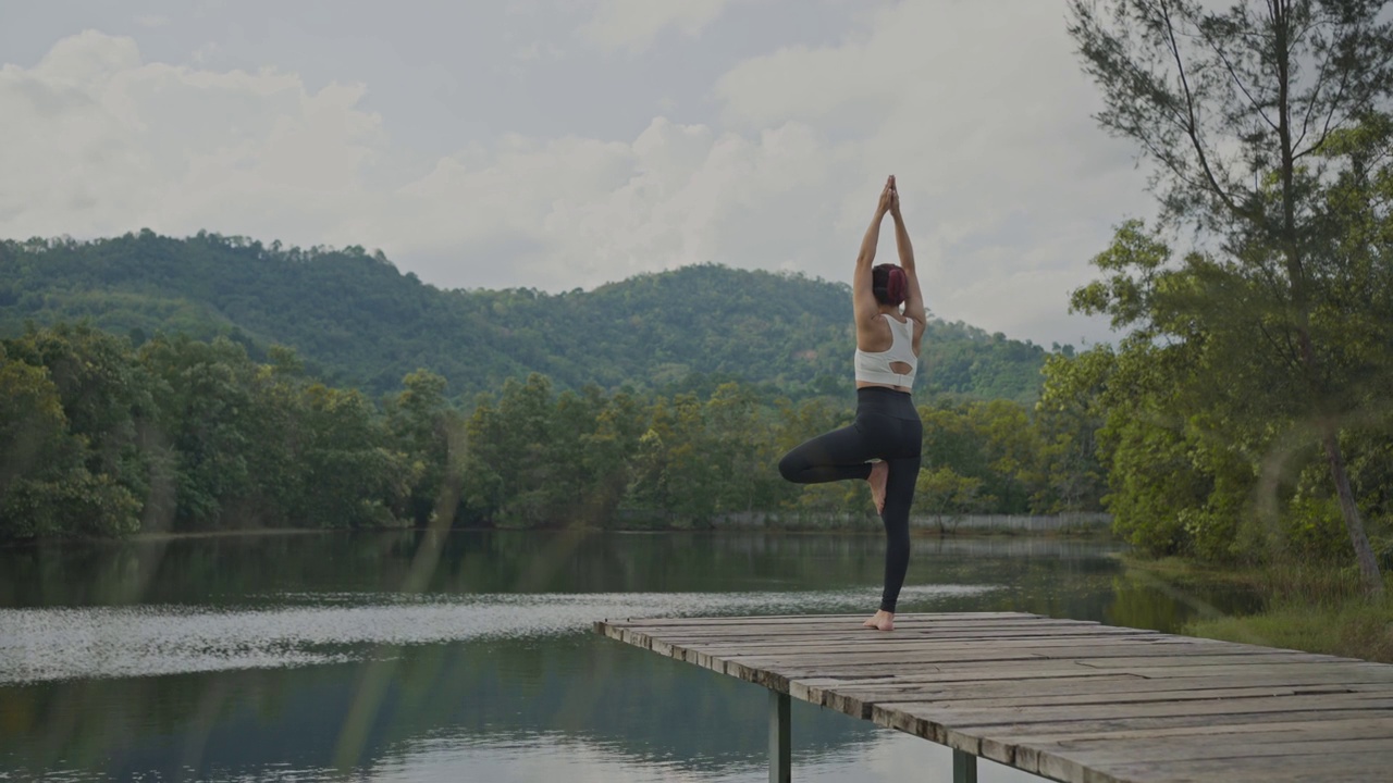 一名亚洲女子在湖边的桥上练习瑜伽。瑜伽和健康生活理念。视频下载