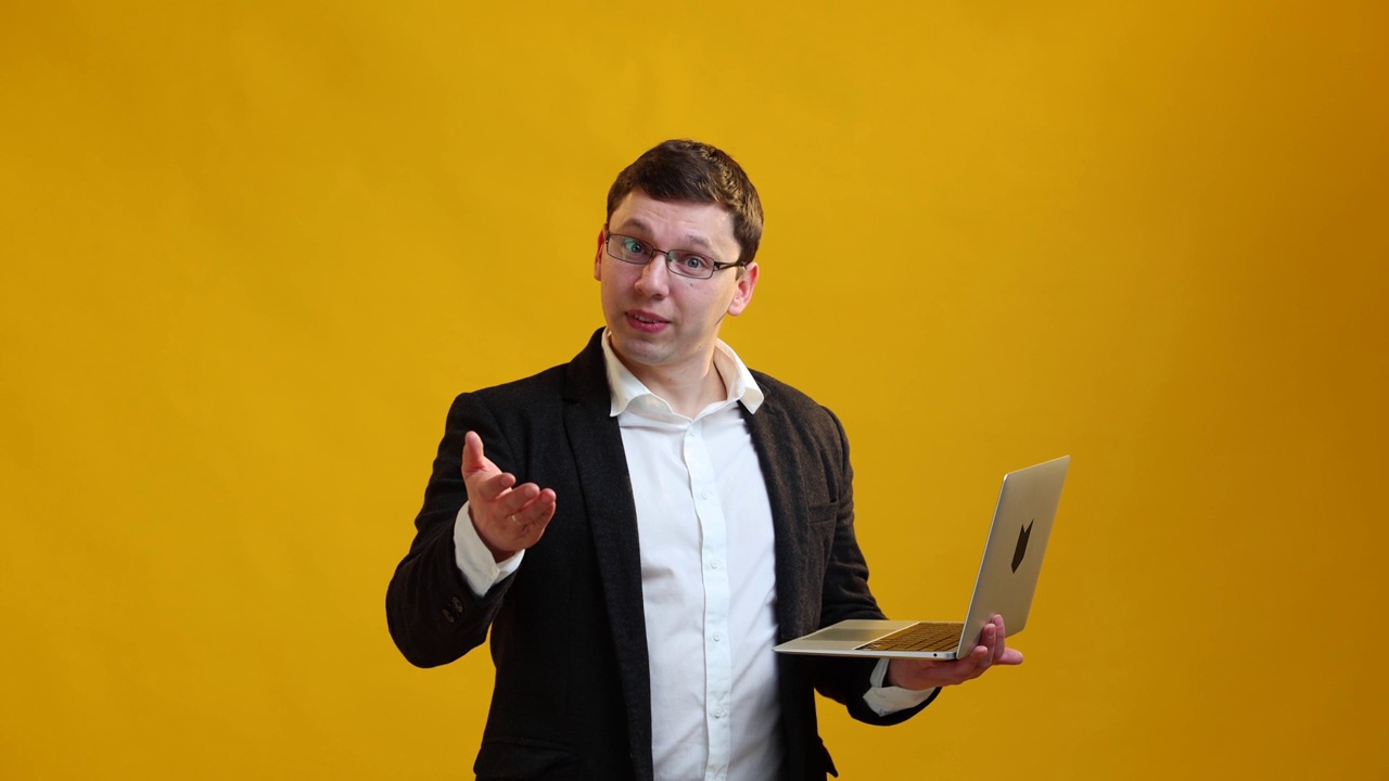 微笑的商人演讲者戴着眼镜在笔记本电脑上显示，在黄色的演播室背景上对着摄像机讲话。经营理念视频下载