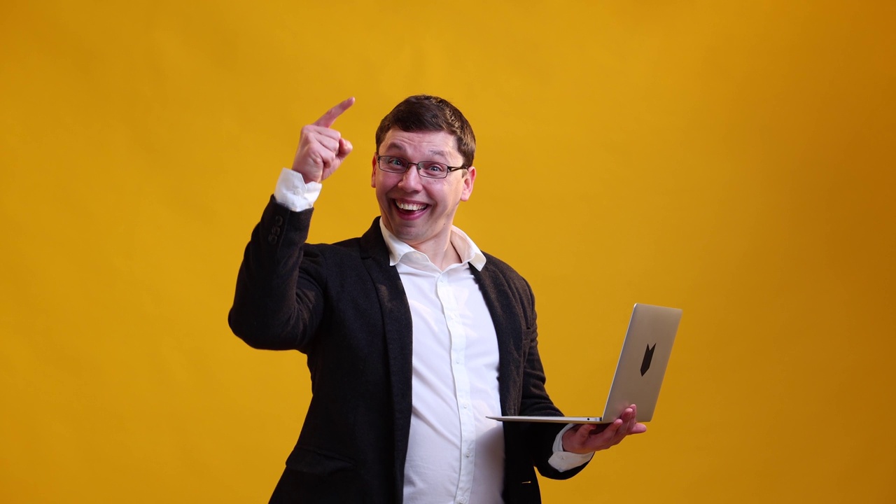 愉快的商人戴着眼镜，展示他的想法，站在黄色的演播室背景上按下笔记本电脑的按钮。线上商业概念视频下载
