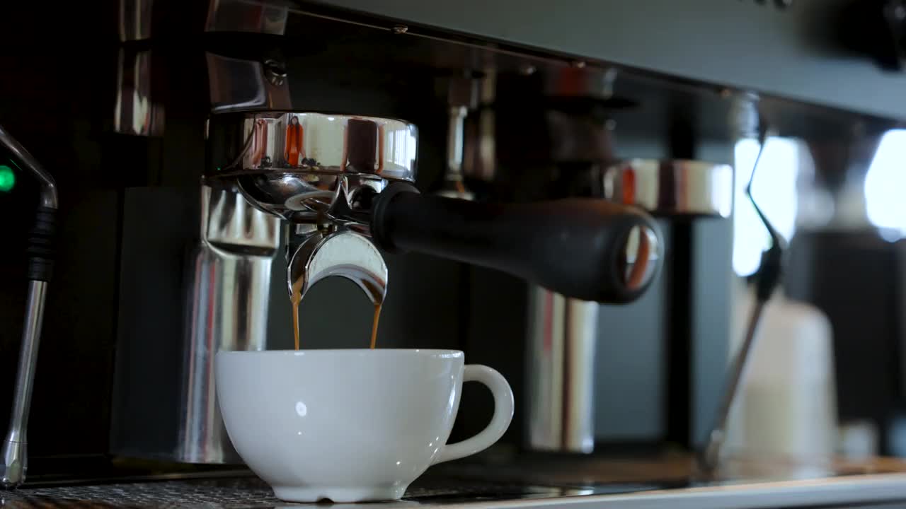 4K，特写白色咖啡杯热菜单，等待咖啡机冲泡的咖啡流入正在等待的咖啡杯，从插入咖啡机的咖啡手柄。视频素材