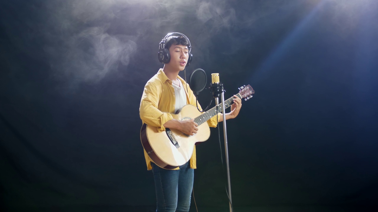 侧视图年轻的亚洲男孩戴着耳机弹吉他，对着电容麦克风在白烟黑色背景上唱歌视频下载