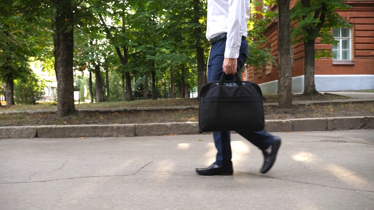 年轻商人带着包的双腿自信地踩在城市街道上。成功交易后，穿着正装的经理或企业家在户外散步。通勤上班的概念。慢镜头侧视图视频下载