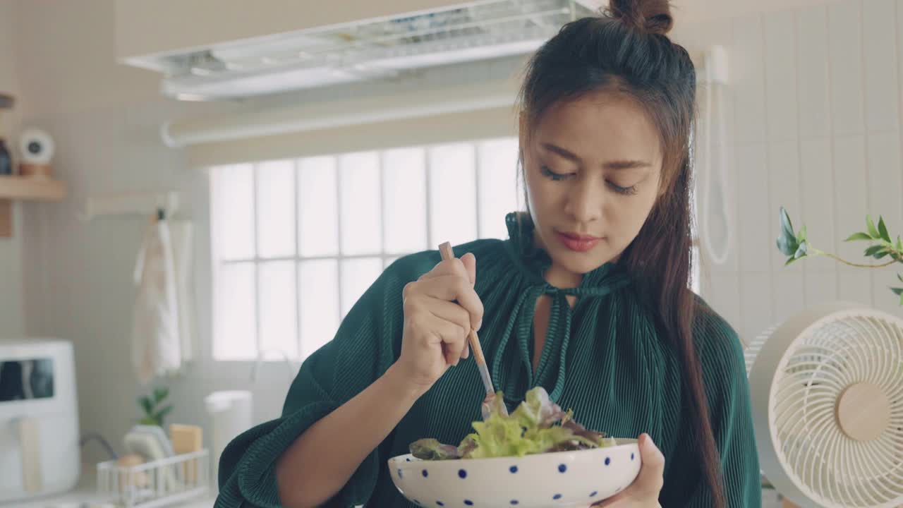 一名亚洲妇女站在厨房里一边用笔记本电脑一边吃沙拉。视频下载