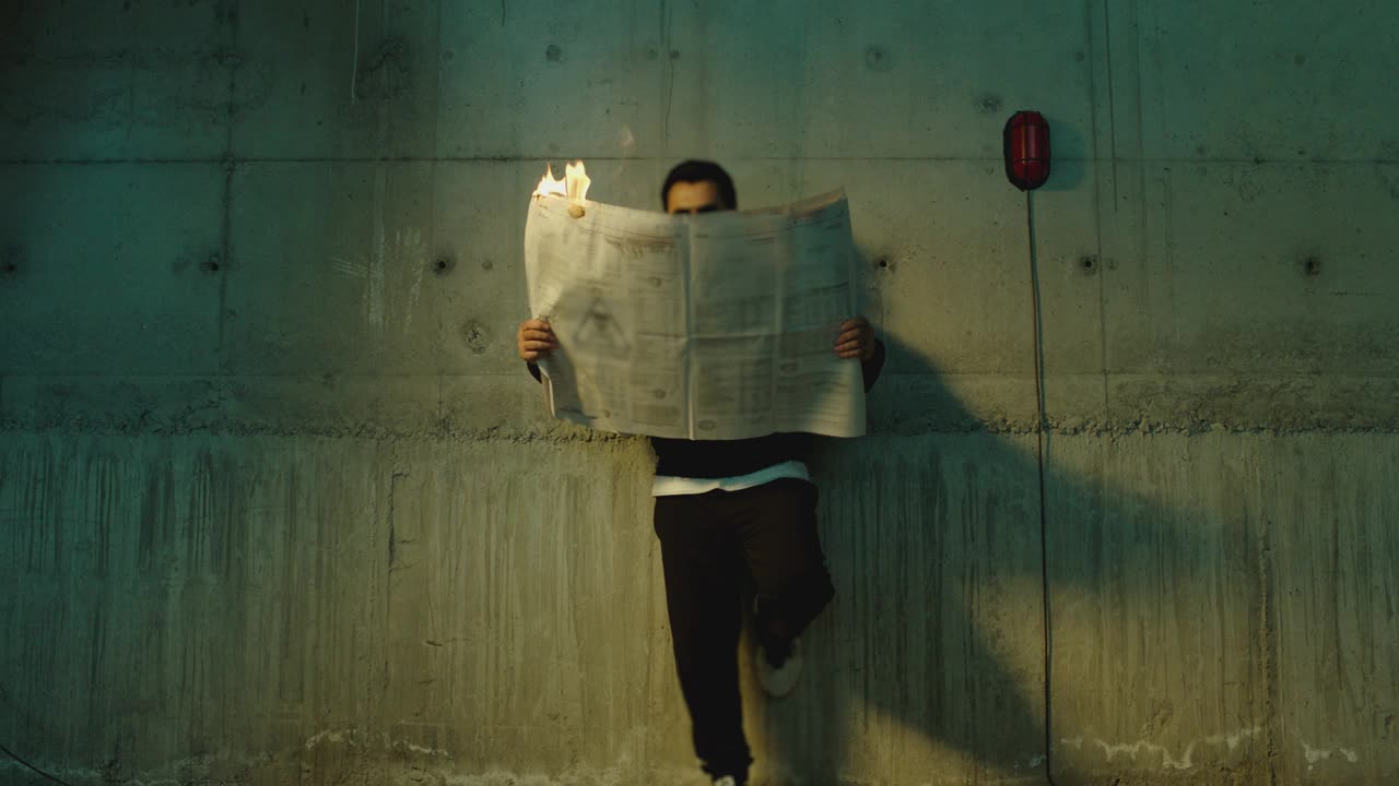 镜头中，男艺术家戴着墨镜站在墙边，在燃烧期间拿着报纸阅读。虚假或禁止新闻的概念或象征。男子阅读燃烧杂志。慢动作视频下载