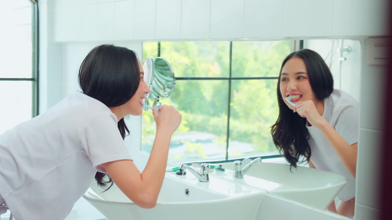 亚洲年轻女子在家里的浴室里刷牙和照镜子。迷人的女性用牙刷和牙膏刷牙，独自享受早晨的口腔防蛀卫生程序。视频下载