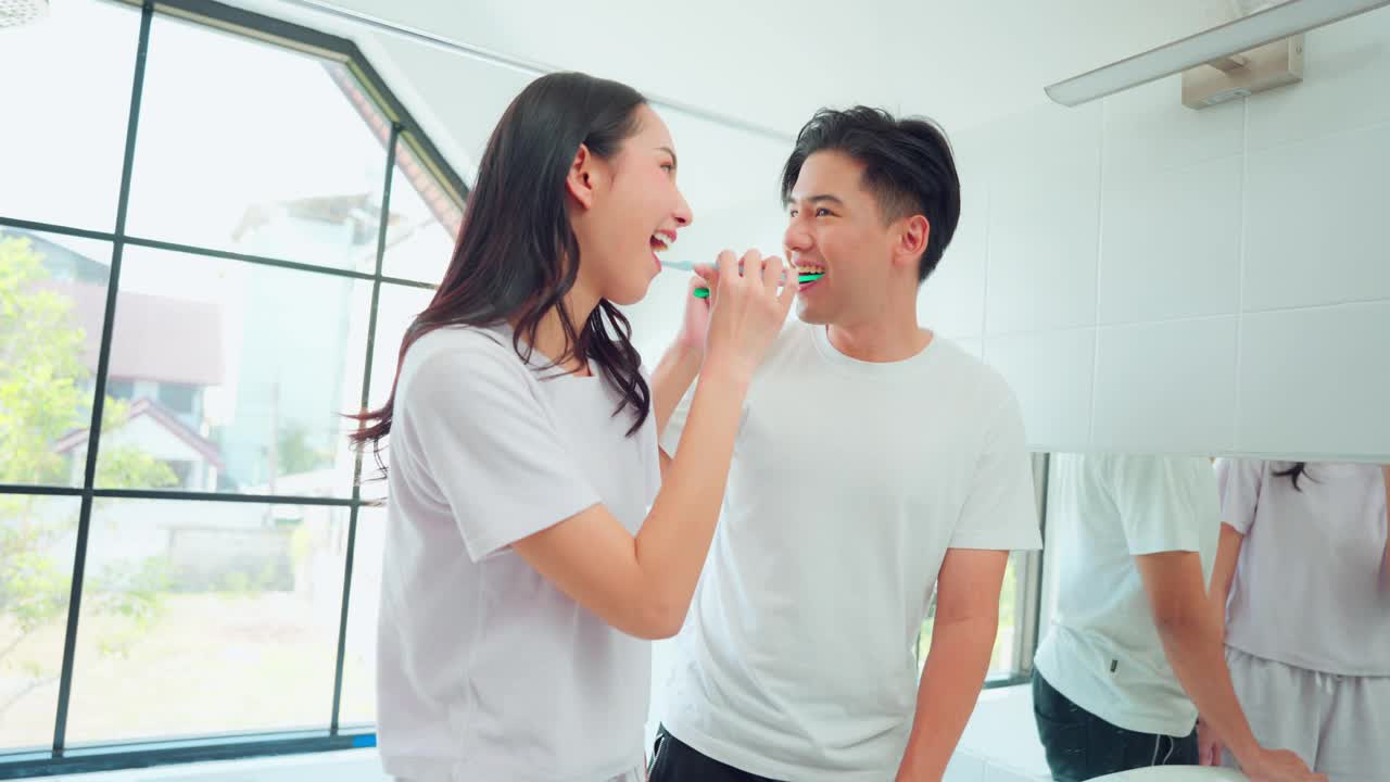 亚洲新婚夫妇在家里的浴室一起刷牙。有魅力的年轻男人和女人感到快乐和放松，享受假期蜜月纪念日，然后看着对方在房子里。视频下载