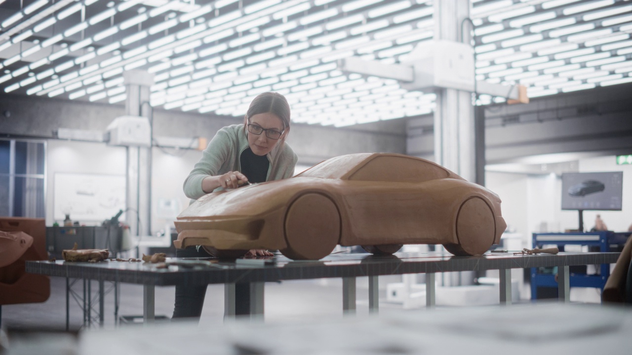 女汽车设计师检查蓝图，用抹刀在模型粘土的原型车上工作。在汽车生产厂的研发工作室工作的建模师视频素材