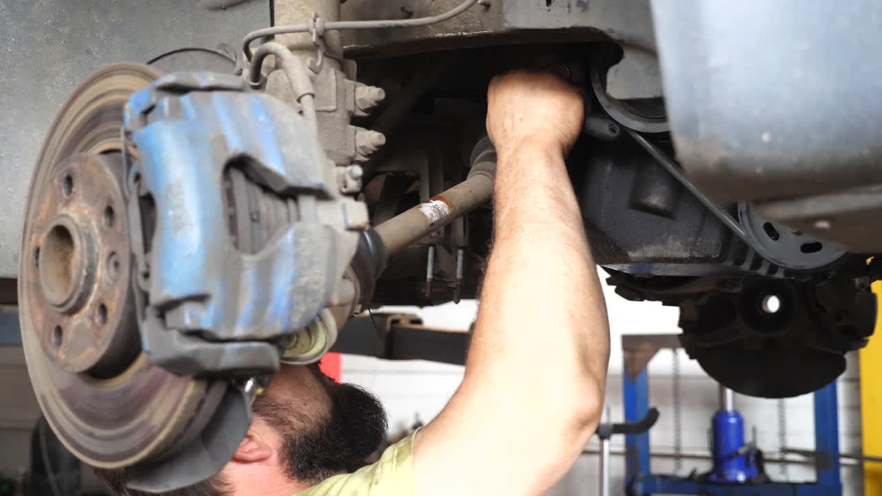 在汽车维修站，不认识的机械师用扳手工具修理汽车。穿着制服的专业修理工在车库的起重车辆下面工作。汽车维修的概念。近距离视频下载