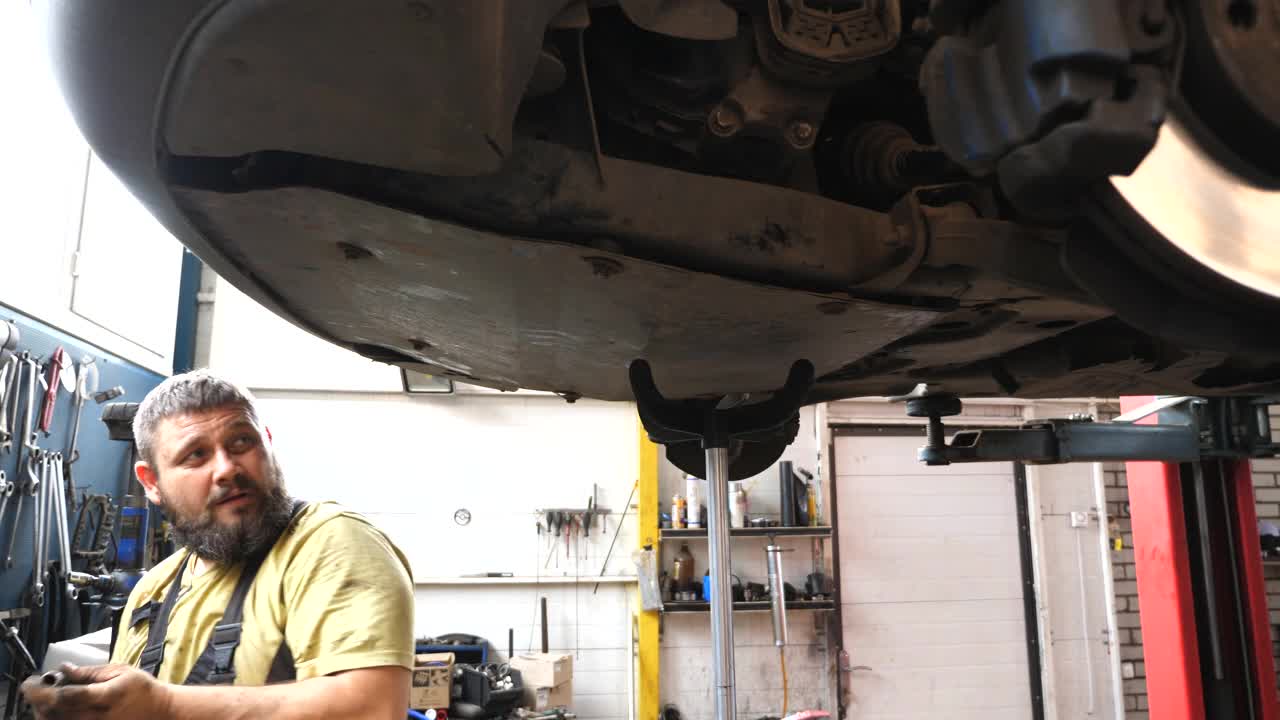 专业汽车技工在车间用专用电动工具紧固螺栓。在车库起重车辆下面工作的工人。大胡子修理工正在修车。汽车维修概念视频下载