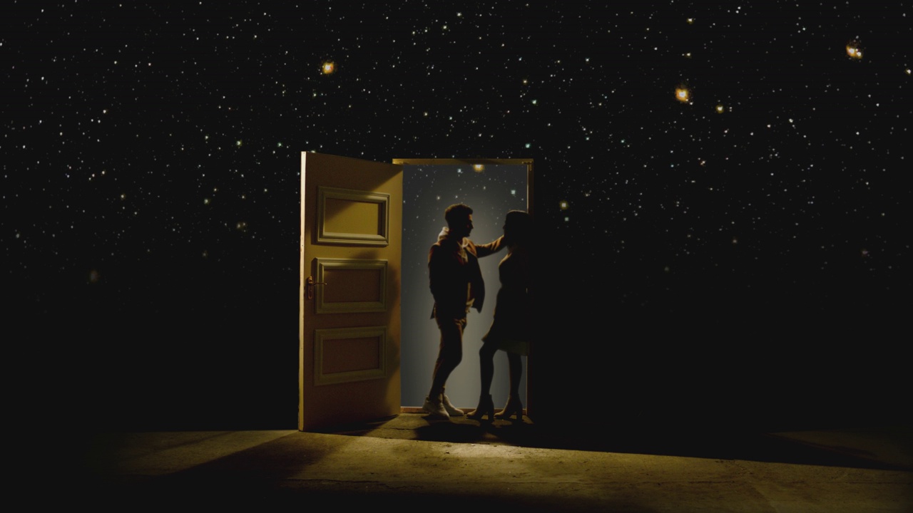 4K侧面视图的夫妇站在对面，看着对方在一扇敞开的门后面的星系。一对情侣站在星星后面的地板上。梦想或浪漫的概念。慢动作视频下载