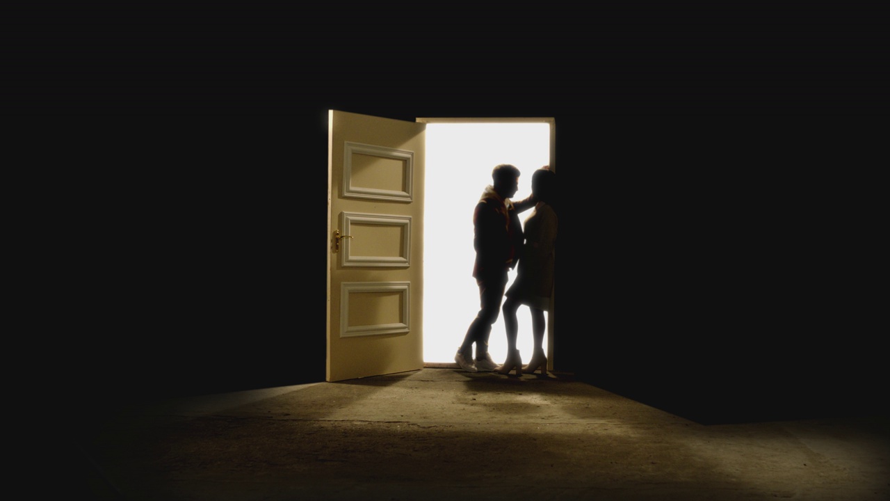 4K侧视图，一对夫妇对面站着，在一扇敞开的门前看着对方，后面有白色的光。一对情侣站在星星后面的地板上。梦幻浪漫的概念。慢动作视频素材