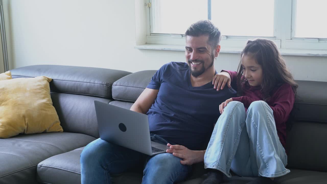 父亲和女儿在客厅的沙发上开心地看着电脑上的互联网内容。视频下载