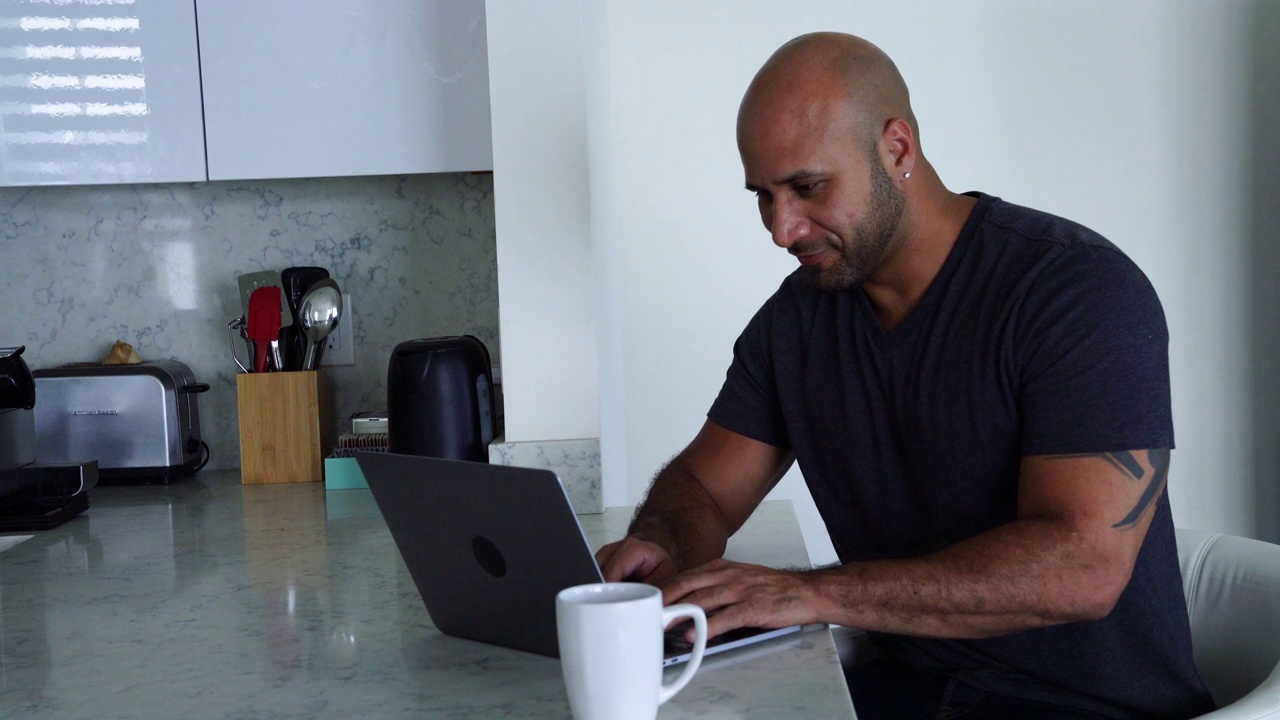 拉丁裔男人一边在厨房喝咖啡，一边愉快地摆弄着他的笔记本电脑视频下载