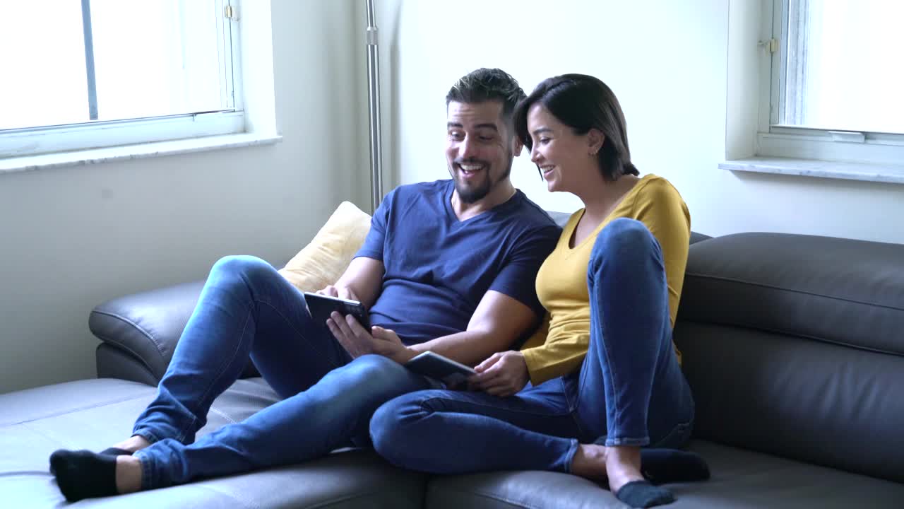 一对中年拉丁裔夫妇在客厅的沙发上通过电子设备观看有趣的内容视频下载