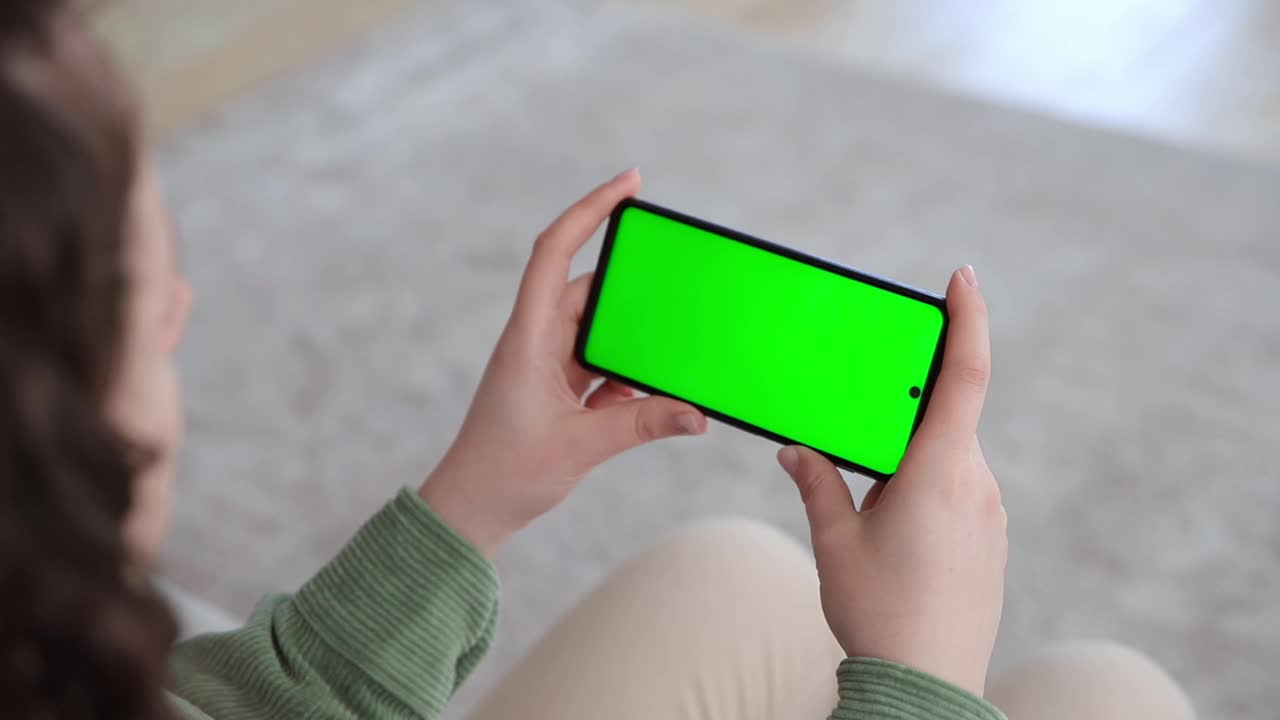 女子使用水平智能手机的背影，屏幕为绿色。彩色键模拟流媒体服务的应用程序。在网上看视频。有选择性的重点视频下载