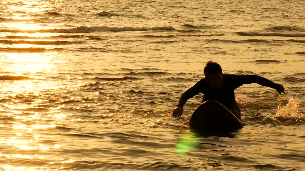 在日落的热带海滩上，冲浪男子划在长长的冲浪板上的剪影视频下载