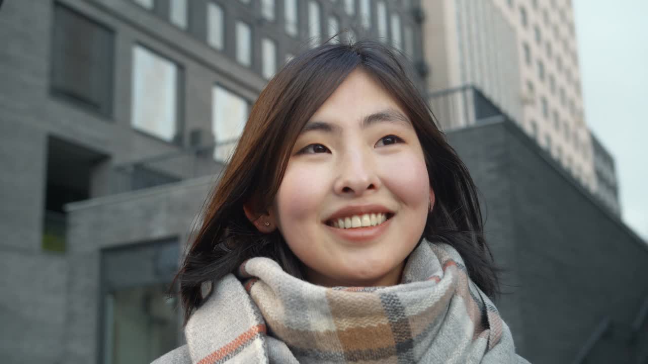 一个亚洲女孩在城市建筑的背景下快乐地微笑的特写。一个黑发的年轻女孩，直视着镜头。自然美景。视频下载
