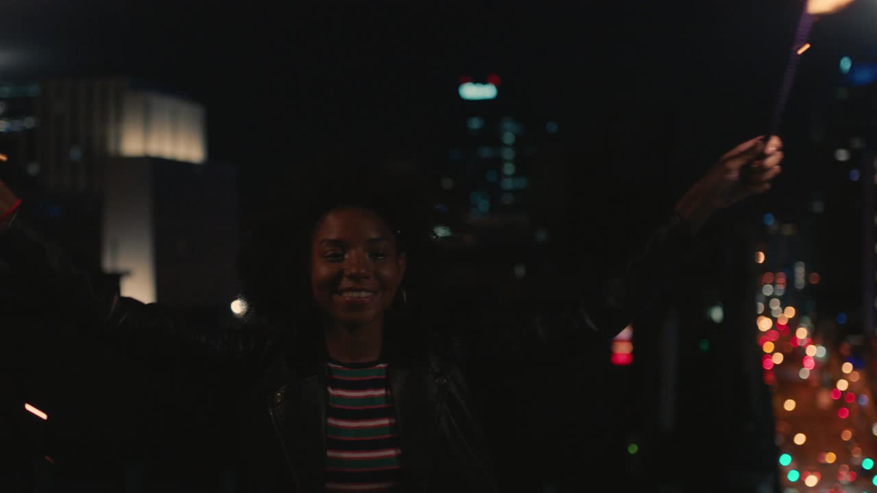 快乐的年轻非裔美国妇女拿着烟花，晚上在屋顶上跳舞，庆祝新年除夕，享受节日庆祝活动视频素材