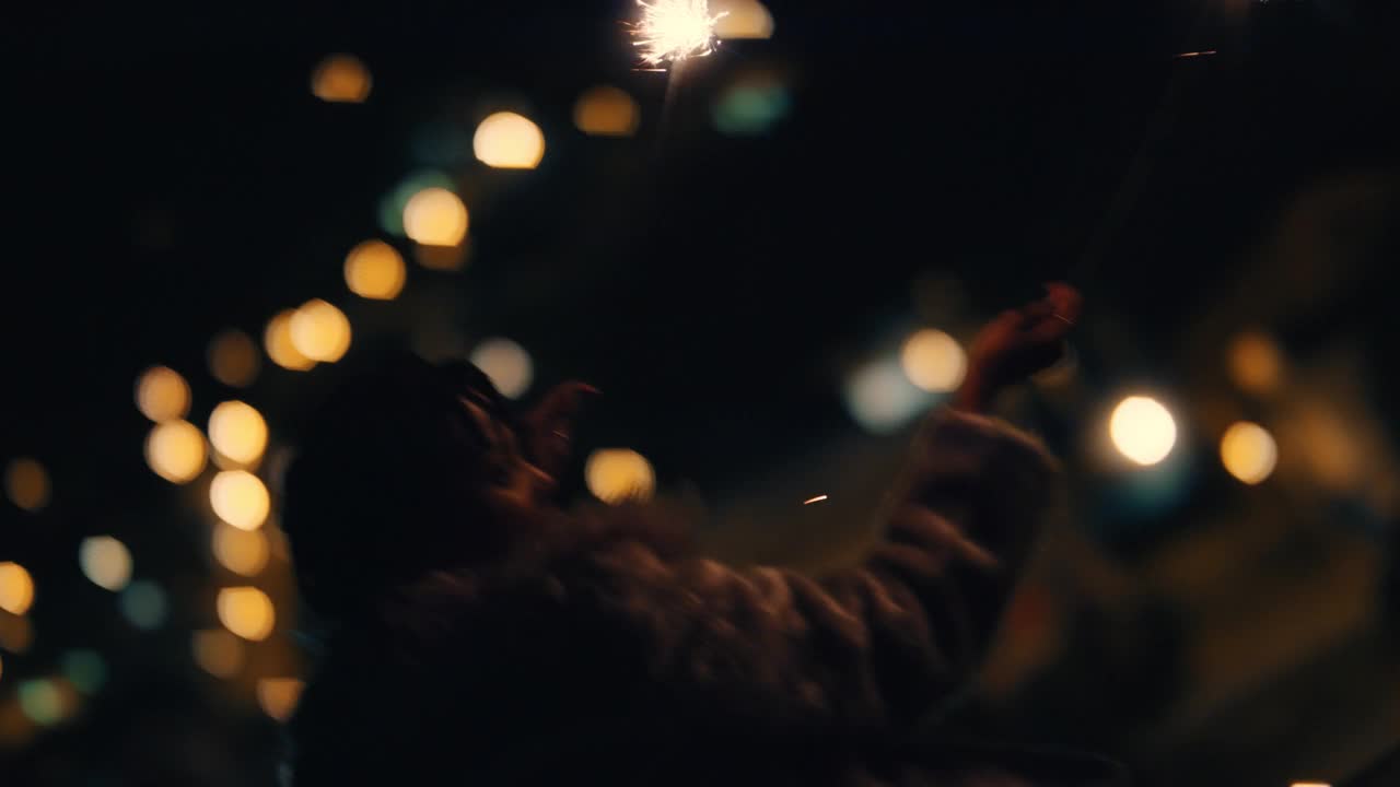 快乐的女人拿着烟花，晚上在屋顶上开玩笑地挥舞着手臂，庆祝除夕，享受独立日庆祝活动，背景是城市的灯光视频素材