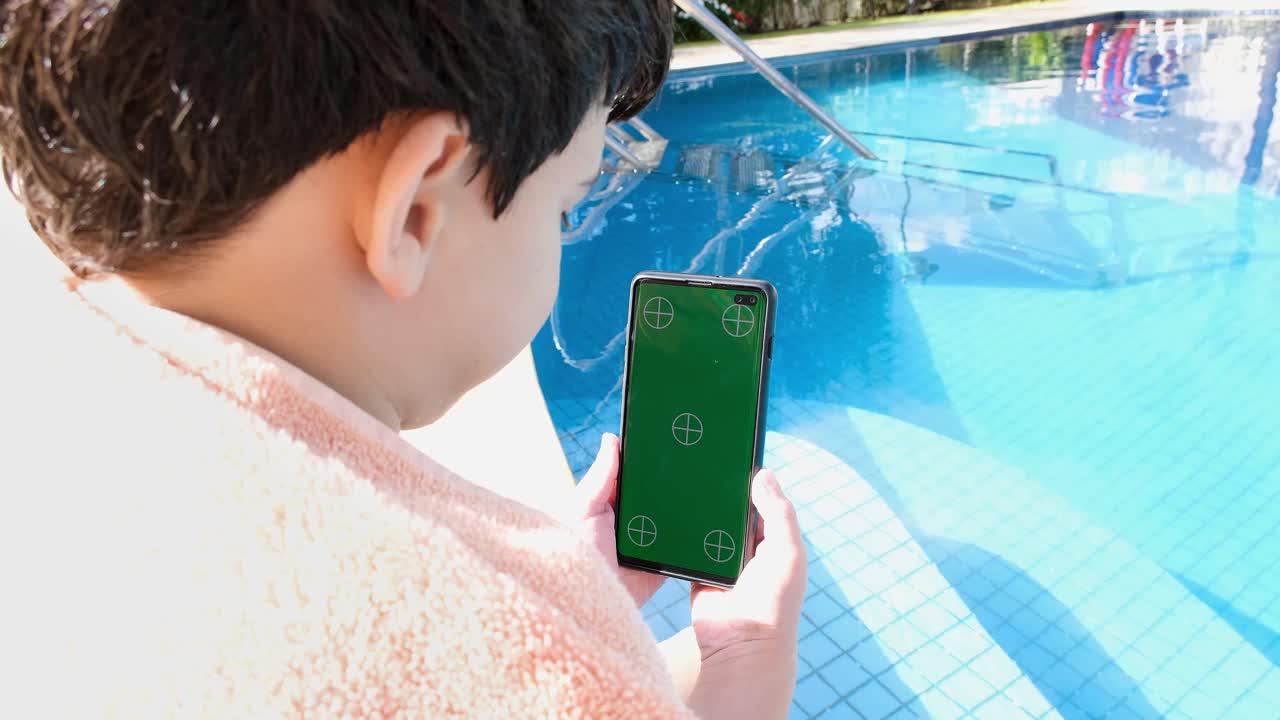 9岁的巴西男孩坐在泳池边，看绿屏的智能手机。视频下载