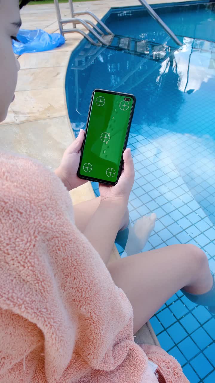 9岁的巴西男孩坐在泳池边，看着绿色屏幕的智能手机。视频下载