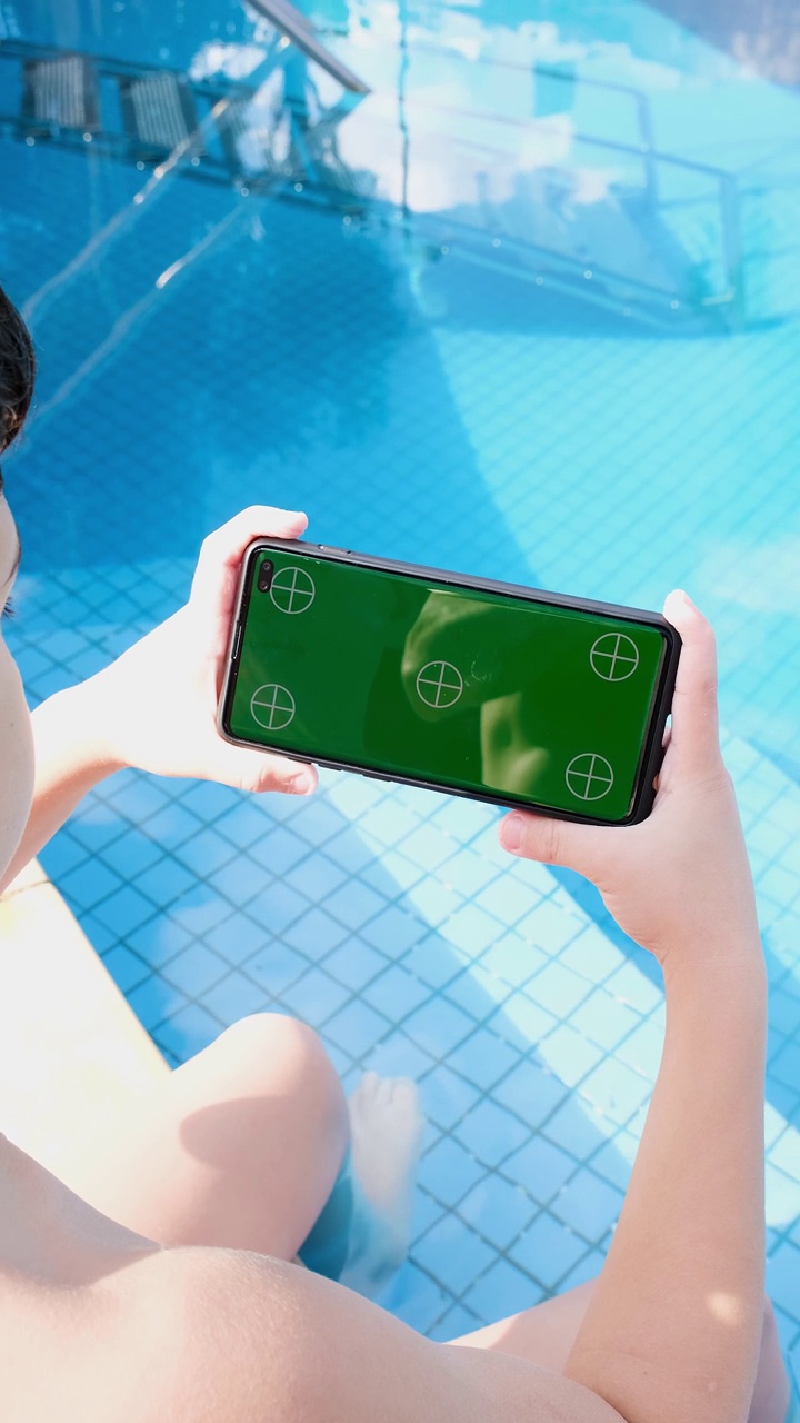 9岁的孩子坐在游泳池里，横着看绿屏的智能手机。视频素材