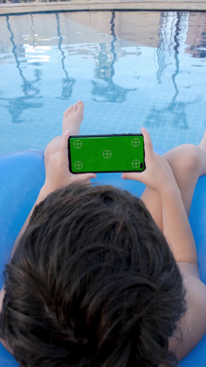 9岁的孩子坐在浮标上，看绿色屏幕的智能手机。视频素材