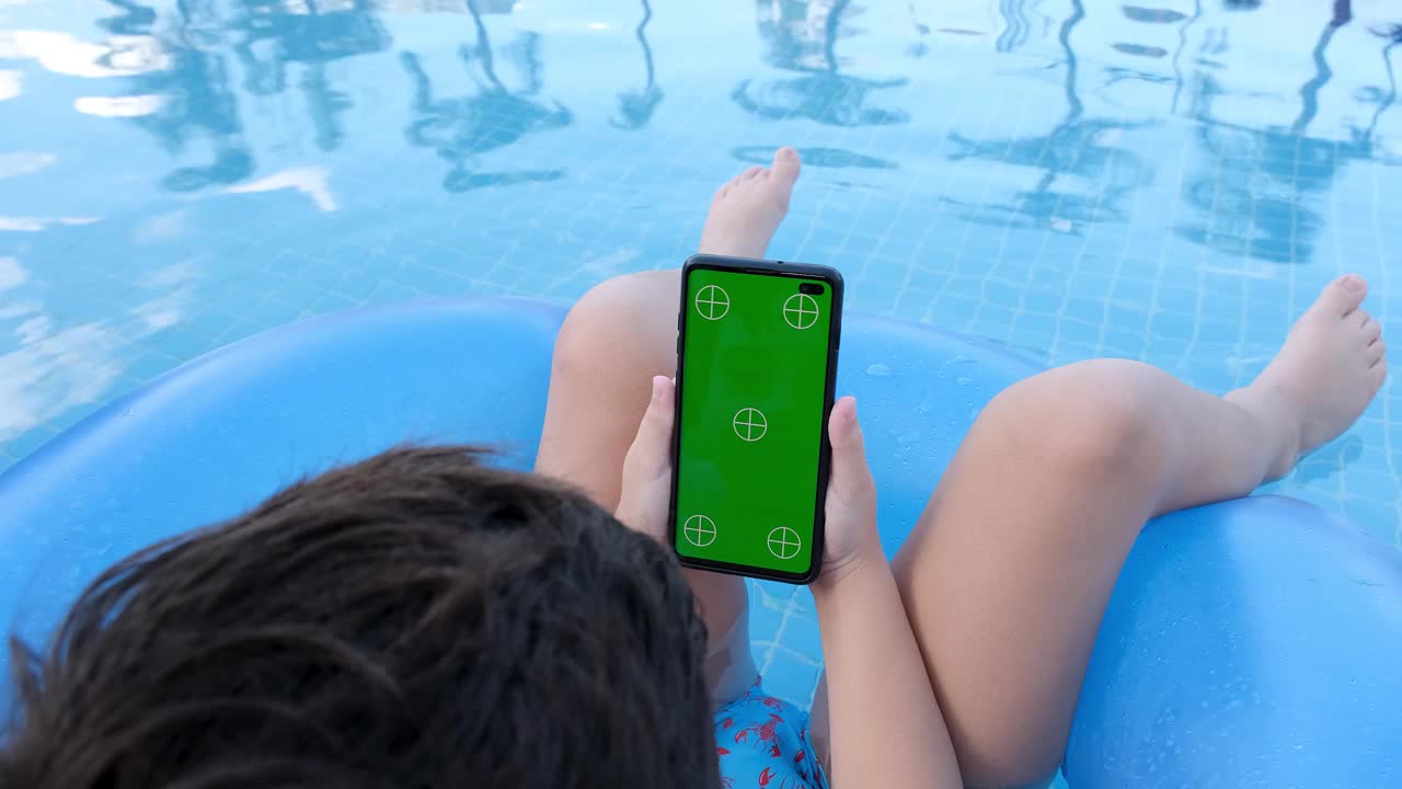 9岁的孩子坐在泳池的浮标上，看着绿屏的智能手机。视频素材