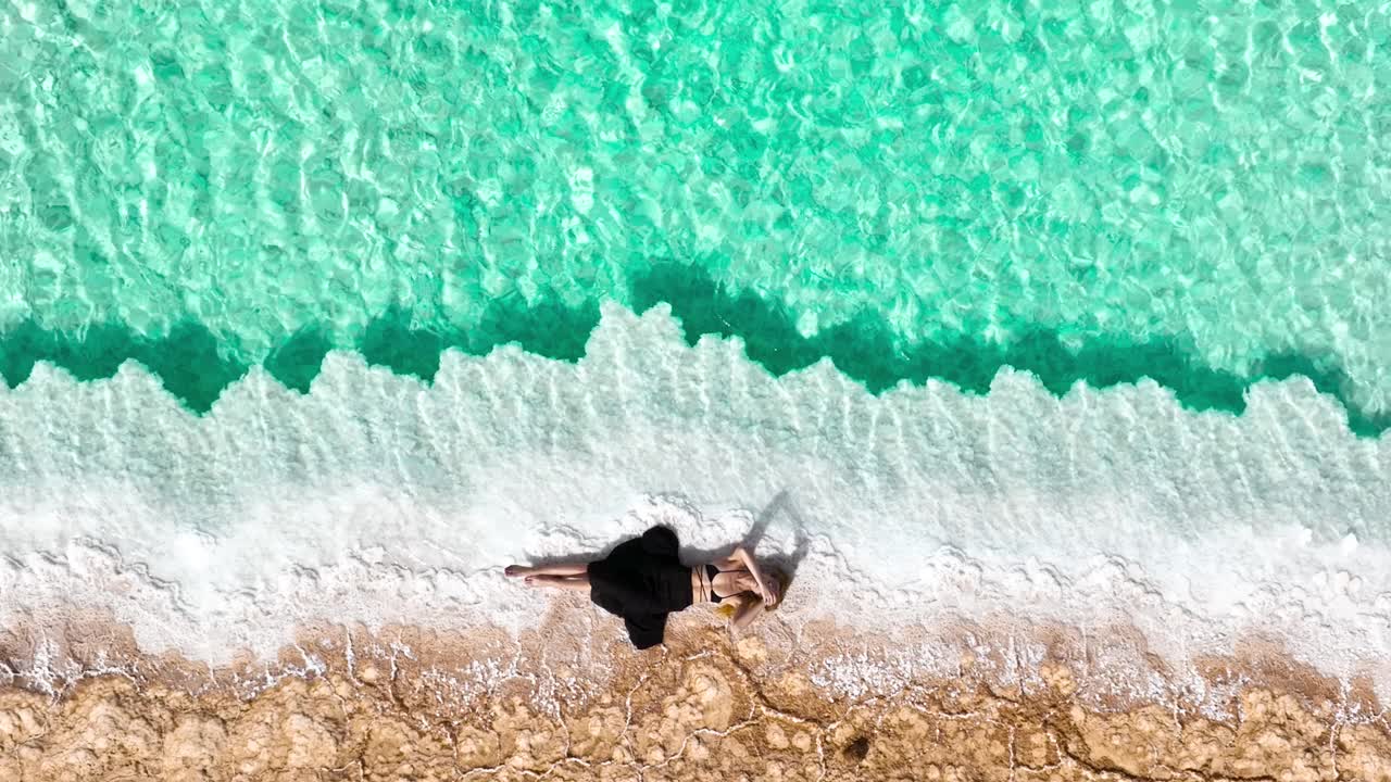 无人机拍摄的埃及锡瓦绿洲，一名年轻女子在绿松石色的盐水池旁晒日光浴的视频。视频下载