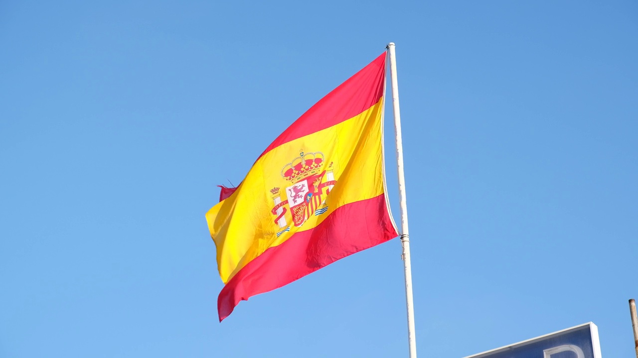 西班牙国旗在蓝天背景上飘扬。旗杆上的旗帜迎风飘扬。红黄相间的西班牙国旗在蓝天上迎风飘扬视频素材