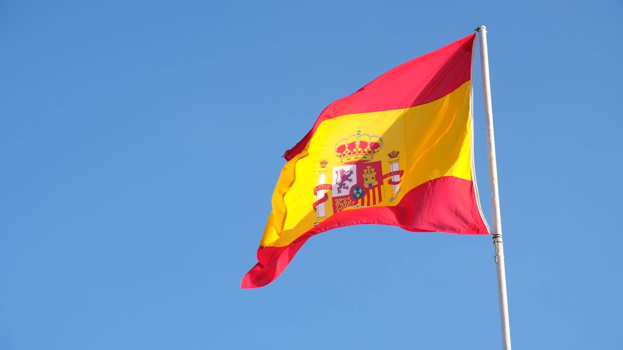 西班牙国旗在蓝天背景上飘扬。旗杆上的旗帜迎风飘扬。红黄相间的西班牙国旗在蓝天上迎风飘扬视频素材