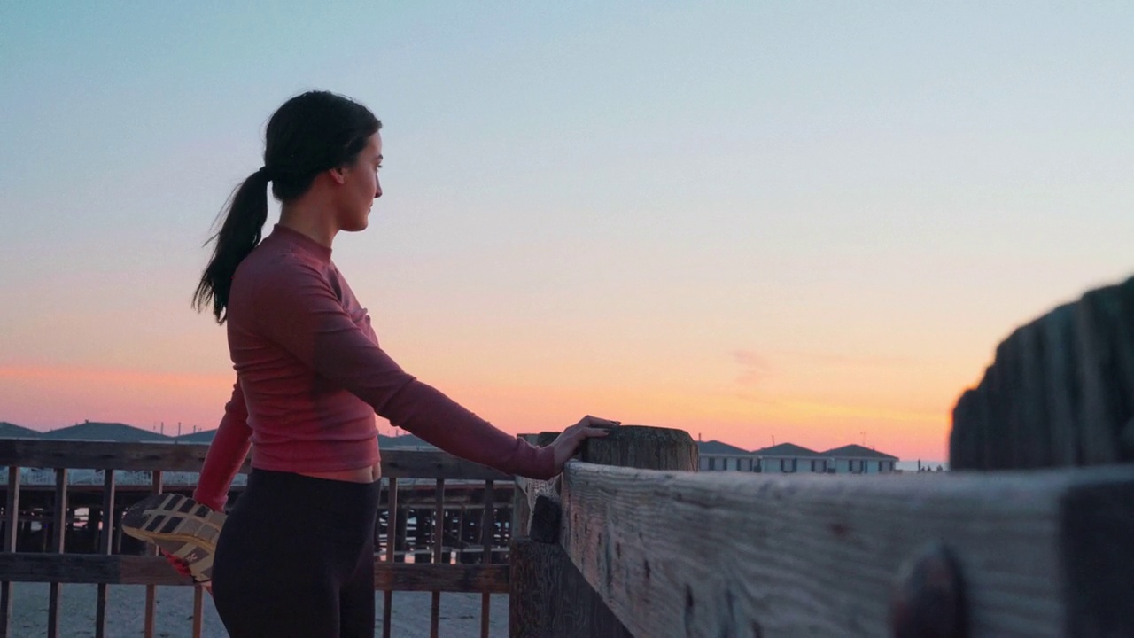 在加州圣地亚哥太平洋海滩的水晶码头上，一位年轻的慢跑女子在日落时分伸展身体，目光远离镜头视频下载