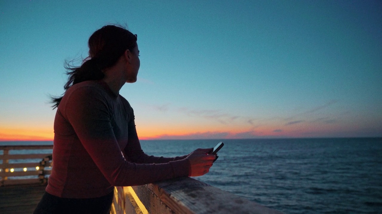 在加州圣地亚哥太平洋海滩的水晶码头，一位年轻的慢跑女性用手机跟踪她的锻炼进度或查看社交媒体应用程序的特写镜头视频下载