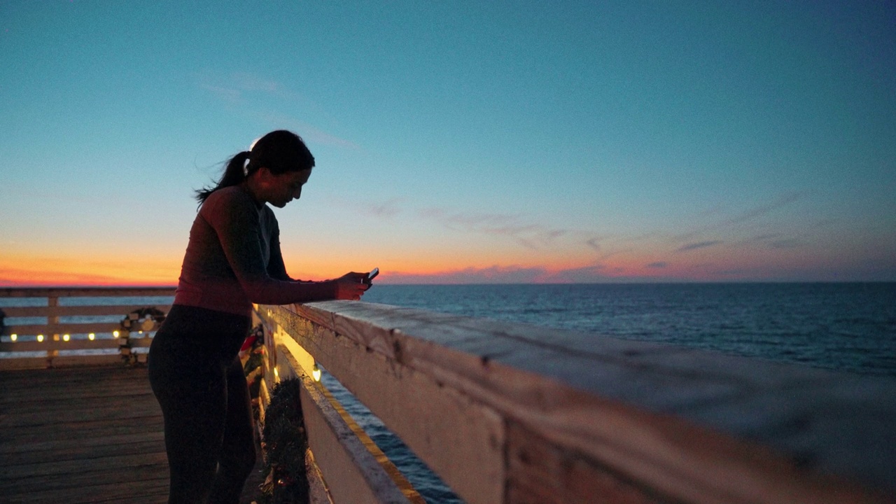 在加州圣地亚哥太平洋海滩的水晶码头，一位沉思、运动的年轻慢跑女性正在休息，用手机跟踪她的锻炼进度或使用社交媒体应用程序视频下载