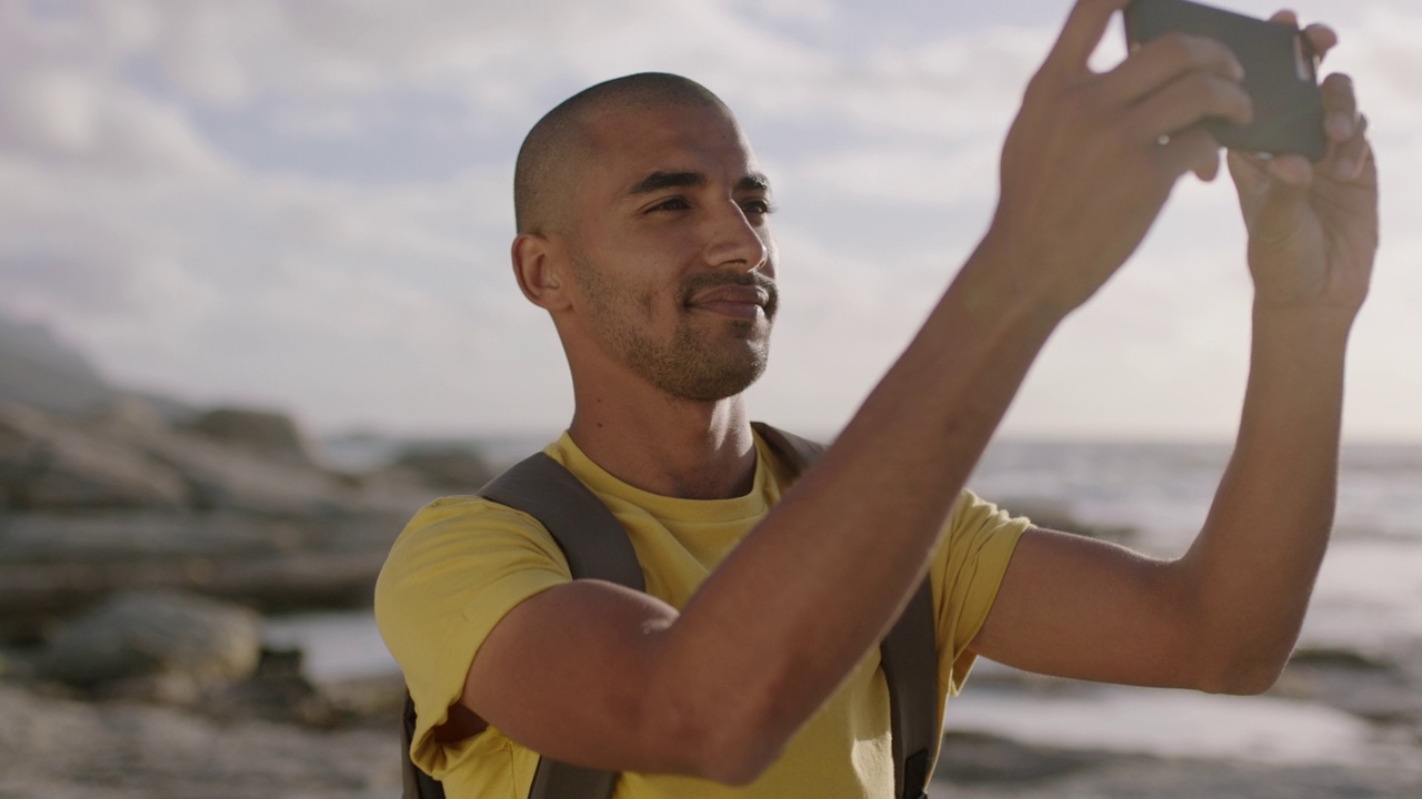年轻迷人的西班牙男子在海滩上用手机拍照，微笑着开心视频素材